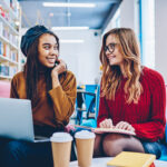 Smiling female students sitting with modern laptop computer and textbook for education at university library, multicultural successful hipster girls communicating indoors during e learning together